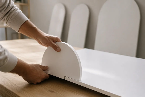 Mani che infilano un piede a mezzaluna nella base di un pannello scenografico bianco appoggiato su un tavolo da lavoro